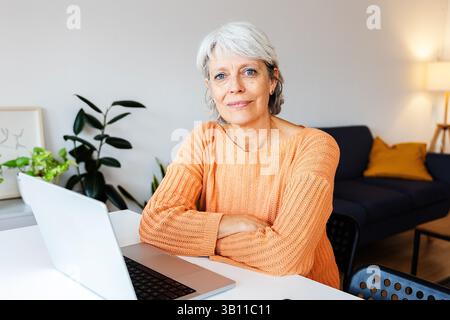 Ältere Frau mit Laptop, die zu Hause auf dem Tisch sitzt Stockfoto