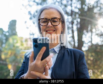 Grauhaarige Geschäftsfrau mit Brille lächelt, während sie im Park ein Smartphone benutzt und die Mischung aus moderner Technologie und Natur genießt Stockfoto
