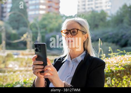 Ältere Geschäftsfrau mit grauem Haar, Brille und formellem Anzug mit Smartphone in einem Stadtpark, Sonnenlicht und Technologie während der Arbeit b Stockfoto