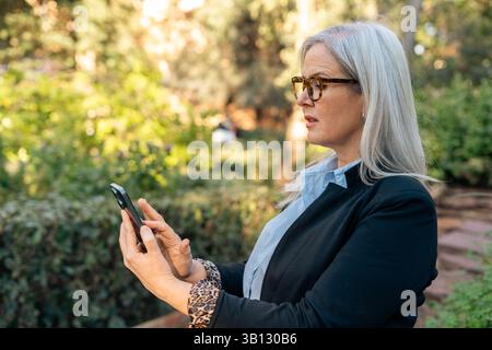 Eine selbstbewusste Profi-Frau, die ihr Smartphone selbstbewusst nutzt, während sie draußen in einer lebendigen und üppigen Gartenumgebung ist Stockfoto