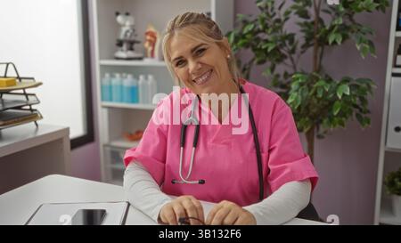 Frau lächelt mit rosa Peelings im Krankenhausbüro mit Stethoskop, während sie am Schreibtisch sitzt und eine professionelle Gesundheitseinrichtung mit Organisation präsentiert Stockfoto