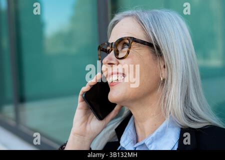 Glückliche, reife Geschäftsfrau mit grauem Haar, die eine Brille trägt, spricht auf dem Handy, während sie in der Nähe eines zeitgenössischen Glasgebäudes steht und angenehm ist Stockfoto