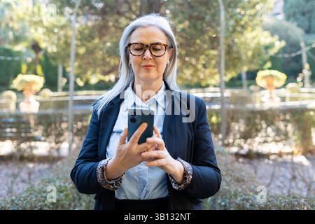 Eine selbstbewusste und reife Frau, die eine stilvolle Brille trägt, greift in einem wunderschönen, üppigen Garten gerne mit ihrem Smartphone in Berührung Stockfoto
