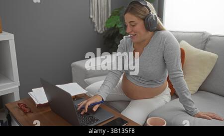 Schwangere Frau mit blonden Haaren arbeitet an einem Laptop in einem gemütlichen Wohnzimmer, trägt Kopfhörer und sitzt bequem auf einem Sofa und veranschaulicht eine ruhige h Stockfoto