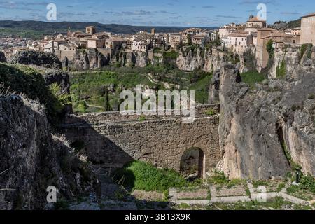 Eine alte Schuttsteinmauer, einst Teil der Grundstücksgrenze des Dichters Federico Muelas, verläuft im Vordergrund vor der Felswand und der Altstadt von Cuenca, Castilla-La Mancha, Spanien. Ein schmiedeeisernes Tor markiert einen Durchgang am Fuße der Mauer, und ein schmaler Pfad führt an der felsigen Nische vom vorherigen Motiv vorbei. Stockfoto