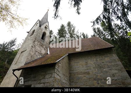 Bad Illstern, italien, verlor seinen Platz. Niedrigwinkelaufnahme einer Steinkirche mit Glockenturm und gekacheltem Dach, umgeben von Bäumen vor einem bewölkten Himmel. Stockfoto