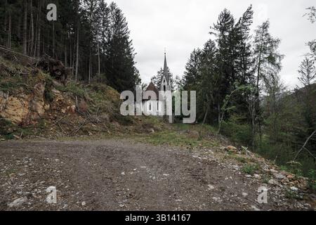 Bad Illstern, italien, verlor seinen Platz. Eine Steinkirche mit einem Kirchturm liegt eingebettet zwischen hohen Bäumen, von einem unbefestigten Pfad unter einem bewölkten Himmel aus gesehen. Stockfoto