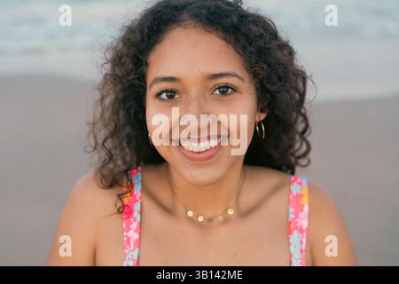 Fröhliche junge Frau mit lockigen Haaren in einem bunten Oberteil, die am Strand warm lächelt. Die ruhige Meereskulisse verstärkt das fröhliche und entspannte Ambiente Stockfoto