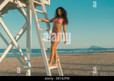 Lächelnde Frau in einem bunten Badeanzug, die auf einer Leiter am Meer steht. Der klare blaue Himmel und der ferne Berg bilden einen ruhigen Strand Stockfoto