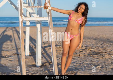 Eine junge Frau in einem lebendigen Badeanzug lehnt sich an einen Rettungsschirm-Turm an einem Sandstrand. Die Meereswellen Rollen sanft unter einem klaren blauen Himmel ein und fangen ein Stockfoto