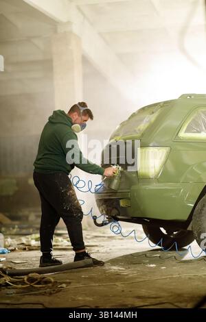 Ein Mann trägt eine Maske, während er ein Fahrzeug in einer schwach beleuchteten Werkstatt mit Staub und Ablagerungen besprüht. Er konzentriert sich auf das Heck des Autos, in A Stockfoto