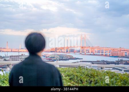 Ein Japaner in seinen Zwanzigern, der eine schwarze Jacke trägt, ist in Minato Mirai, Yokohama, Präfektur Kanagawa. Von hinten gesehen beobachtet er eine ferne Brücke Stockfoto