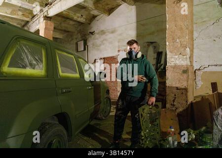 In einer schattigen Garage trägt ein Mann mit einer Gasmaske grüne Farbe auf einen SUV auf und konzentriert sich darauf, sein Erscheinungsbild mit einem lebendigen Design in einer Hand-o-Form zu verändern Stockfoto