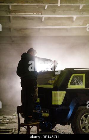 Ein Mann steht auf einem Stuhl und besprüht ein Auto in einer schwach beleuchteten Garage. Die Atmosphäre ist mit Nebel gefüllt, was die fokussierte Arbeit am Fahrzeug unterstreicht. Stockfoto