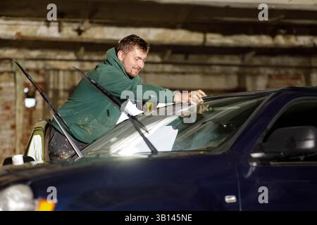 Ein Mann repariert sorgfältig eine Windschutzscheibe in einer Garage, wobei er sich auf Präzision konzentriert. Die Einstellung ist schwach beleuchtet, mit sichtbaren Werkzeugen und einem zweiten Fahrzeug im Hintergrund Stockfoto