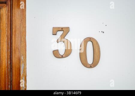Vintage Messing Haus Nummer 30 auf weißer Stuckwand mit rustikalem hölzernem Türrahmen. Klassisches mediterranes Gebäude, Adresszahlen, die Alter und Alter anzeigen Stockfoto