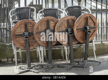 Newcastle, Großbritannien - 13. April 2025 - Holztische und Edelstahlstühle stapeln sich entlang des Eisenzauns neben dem Fußweg vor dem Pub & Restaurant. u Stockfoto