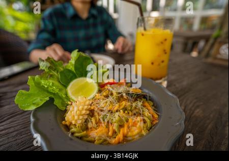 Person genießt Meeresfrüchte-Pasta und grünen Salat mit Limettenkeil auf einem zwanglosen Restauranttisch, mit erfrischendem Passionsfruchtsaft und Highlight Stockfoto