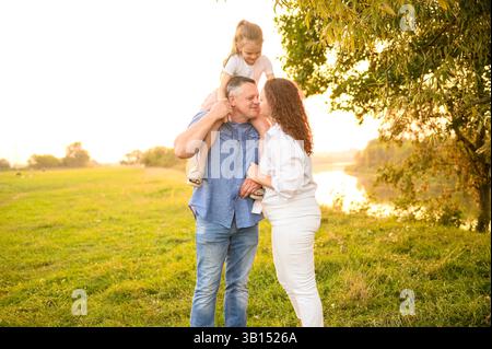 Glückliche Familie bei Sonnenuntergang während des Spaziergangs und der Erholung im Park. Dad hat Tochter am Hals, Mom umarmt ihn, küsst ihn, sieht mit Liebe aus. Wochenende in der Natur, Familienbeziehungen Stockfoto