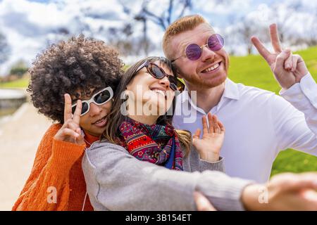 Drei fröhliche, ethnische Freunde, die eine Sonnenbrille tragen und ein Friedenszeichen beim Selfie im Park machen Stockfoto