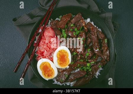 Udon japanisches Rindfleisch mit Reis, eingelegtem Ingwer, gekochter Eierscheibe, garniert mit grünen Zwiebeln, in einer schwarzen Schüssel, mit Stäbchen Stockfoto