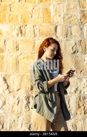Fokussierte junge Frau mit rotem Haar, die vor einer Steinmauer steht und ein Smartphone hält und benutzt, während sie in legeren Kleidern in warmen Außenlichtern gekleidet ist Stockfoto