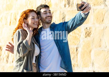 Glückliches junges Paar, das nahe beieinander steht, lächelt und winkt, während es ein Selfie mit einem Smartphone vor einem hellen, sonnendurchfluteten Steinmauerhintergrund macht. Stockfoto