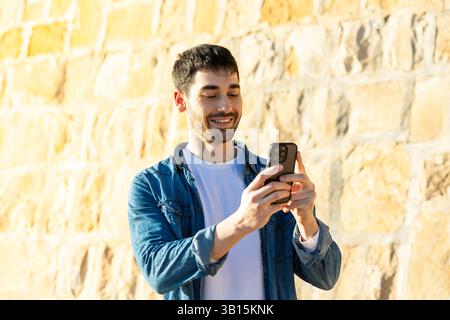 Ein lächelnder junger Mann, der vor einer sonnendurchfluteten Steinmauer steht, während er ein Jeansshirt trägt, auf sein Smartphone blickt und einen hellen Outdoor-Moment genießt Stockfoto