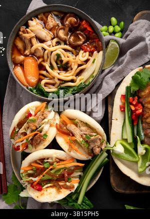 Chinesische Gerichte auf dunklem Hintergrund: Süße und saure Ente, Nudelsuppe, gedämpfte Brötchen. Bankett oder Buffet im asiatischen Stil, Blick von oben. Stockfoto