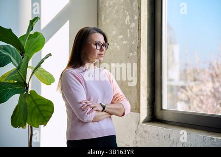 Ruhige, schöne Frau mittleren Alters, die aus dem Fenster blickt Stockfoto