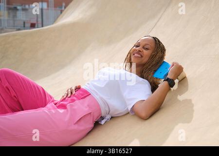 Stilvolle Frau, die sich auf der Skate-Rampe entspannt, Penny-Board hält und lächelt Stockfoto