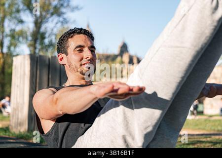 Fitter Mann, der V-Sit-Bauchtraining auf einer Outdoor-Matte durchführt, lächelt und balanciert mit ausgestreckten Armen und Beinen bei hellem Tageslicht im Park Stockfoto