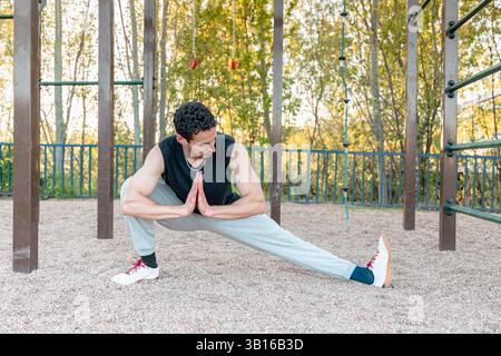 Mann, der im Calisthenics-Park im Freien Kosaken-Hocken-Stretching für Hüftbeweglichkeit, Balance und Beinflexibilität macht, und Fitnesskleidung trägt Stockfoto