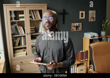 Hochwinkelporträt eines afroamerikanischen Priesters mit Tablette in den Händen, die im Büro zum Foto posiert Stockfoto