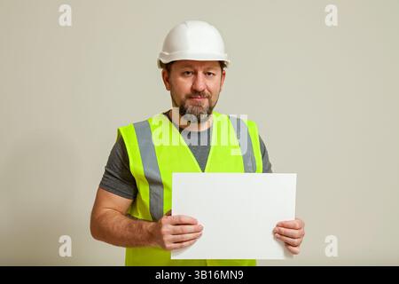 Männlicher Bauarbeiter mit weißem Helm und reflektierender Weste, stehend vor neutralem Hintergrund und hält ein leeres weißes Schild für Kopien Stockfoto