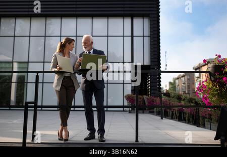 Zwei Geschäftsleute, die gerne Gespräche führen und Dokumente auf Papier und Laptop vor dem Bürogebäude lesen Stockfoto