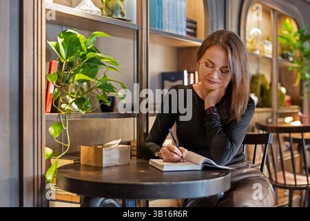 Eine Frau schreibt Notizen an einem stilvollen Kaffeetisch, eine junge Frau mit Brille schreibt in ein Notizbuch in einem Café. Stockfoto