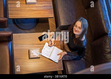 Eine Frau schreibt im Tagebuch am Kaffeetisch. Eine Frau in Brille schaut auf, während sie in einem Tagebuch an einem hölzernen Kaffeetisch schreibt, mit Smartphone und Taschentuch Stockfoto
