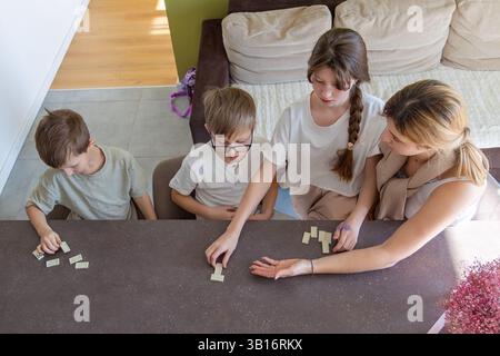 Familie spielt gemeinsam Dominoes zu Hause. Mutter und drei Kinder genießen eine gute Zeit beim Spielen eines Dominospiels an einem Tisch in einem gemütlichen Wohnzimmer Stockfoto