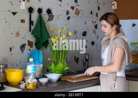 Frau, die Essen in der modernen Küche zubereitet. Eine Frau steht an einer Küchentheke, hält ein Messer und bereitet Essen auf einem Schneidebrett vor, umgeben von Stockfoto
