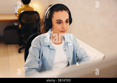 Geschäftsfrau in einem modernen Büro, die Kopfhörer trägt und sich während der Arbeit auf ihren Computerbildschirm konzentriert. Produktivität und Konzentration an einem typischen Tag. Backgr Stockfoto