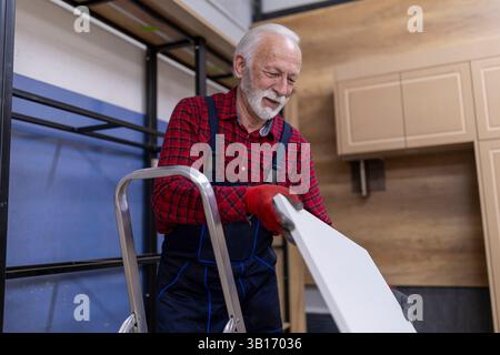 Mann mit grauen Haaren und Bart, der auf einer Leiter steht und in einer Werkstatt Holzregale zusammenbaut, mit Schutzhandschuhen Stockfoto