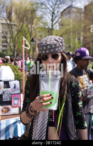 Eine junge Frau mit langen dunklen Haaren, die einen gemusterten Hut, eine Sonnenbrille und eine Jacke mit Cannabisblättern trägt, trinkt aus einem Beutel mit hellgrünem Liqu Stockfoto