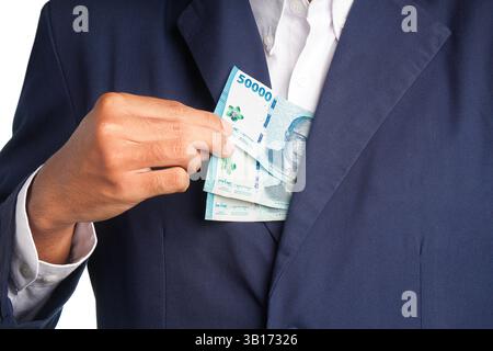 Eine Nahaufnahme, bei der die Hand eines Geschäftsmannes gefangen wurde, der mehrere fünfzigtausend indonesische Rupiah (IDR)-Banknoten in die Innentasche seines blauen s einsteckte Stockfoto