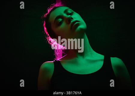 Junge Frau mit erhobenem Kopf posiert unter dramatischem Neonlicht mit rosa Leuchten, die ihre Silhouette vor dunklem Studio-Hintergrund hervorheben. Stockfoto