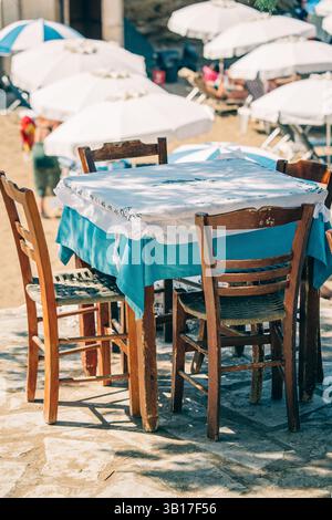 Leerer Holztisch und Stühle in einer Taverne am Meer mit Blick auf einen sonnigen Strand mit verschwommenen Sonnenschirmen und Touristen in Griechenland. Stockfoto