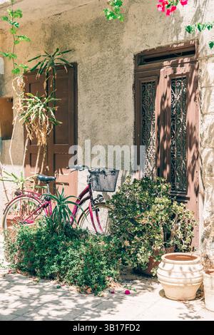 Vor einem traditionellen griechischen Haus auf Kreta, Griechenland, liegt ein rotes Fahrrad vor einer sonnendurchfluteten Wand mit lebhaften Bougainvillea. Stockfoto