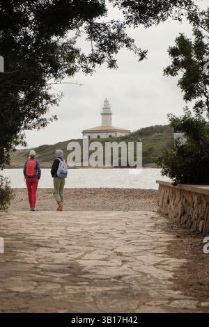 Zwei Touristen gehen auf einem Steinweg zu einem Leuchtturm auf einem Hügel auf Mallorca, eingerahmt von Bäumen. Stockfoto