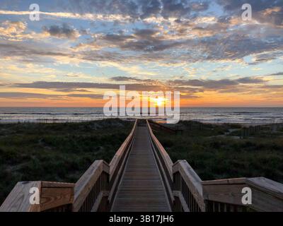 Ein Blick auf eine hölzerne Promenade, die zum Strand führt, wo die Sonne durch die Wolken ragt, farbenfroher Himmel und Ozean im Hintergrund. Amelia Island, Florid Stockfoto