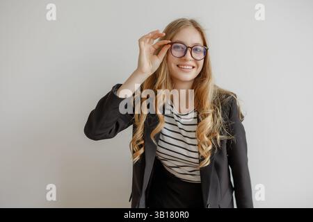 Eine junge Frau mit langen Haaren, die eine Brille und ein gestreiftes Hemd trägt, posiert zuversichtlich in einem schwarzen Blazer drinnen Stockfoto